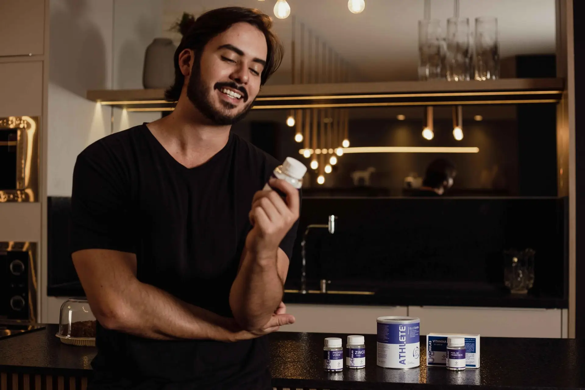 A smiling man in a modern kitchen holds a bottle of vitamins while looking at it. Various supplement bottles, including zinc and other health products, are displayed on the counter in front of him. The scene is warmly lit, emphasizing the importance of men's health and wellness through the use of vitamins and supplements.