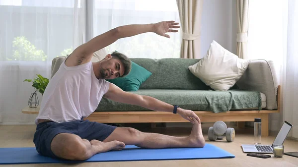 A man stretching on a yoga mat in his living room, reaching toward his foot with one arm extended overhead. This image illustrates the benefits of stretching and highlights the importance of flexibility for overall fitness